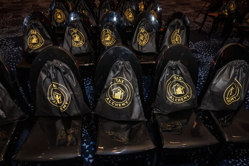 Black drawstring bags with the gold Tax Alchemy logo hanging on rows of chairs