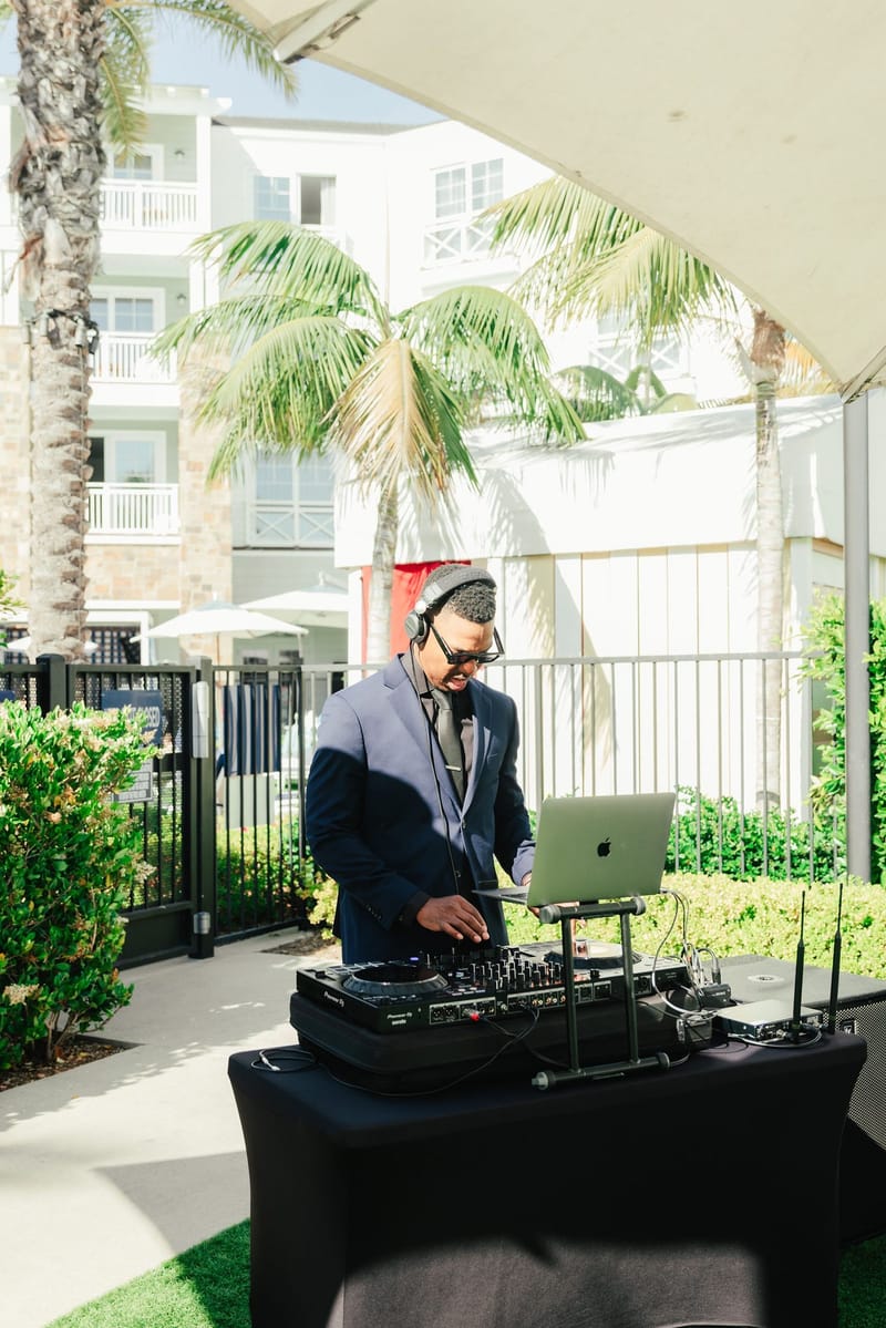 A DJ in headphones at a laptop and decks on a terrace with palm trees behind