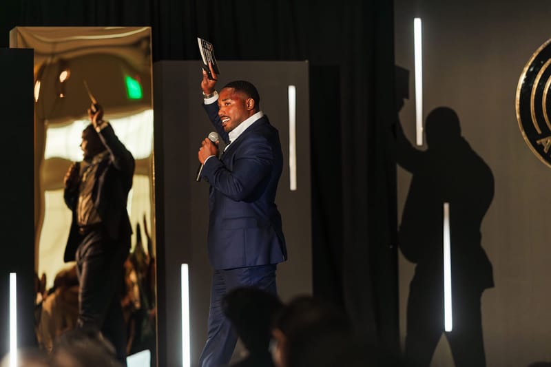 A speaker in a navy suit raising one hand and holding a microphone between vertical light bars