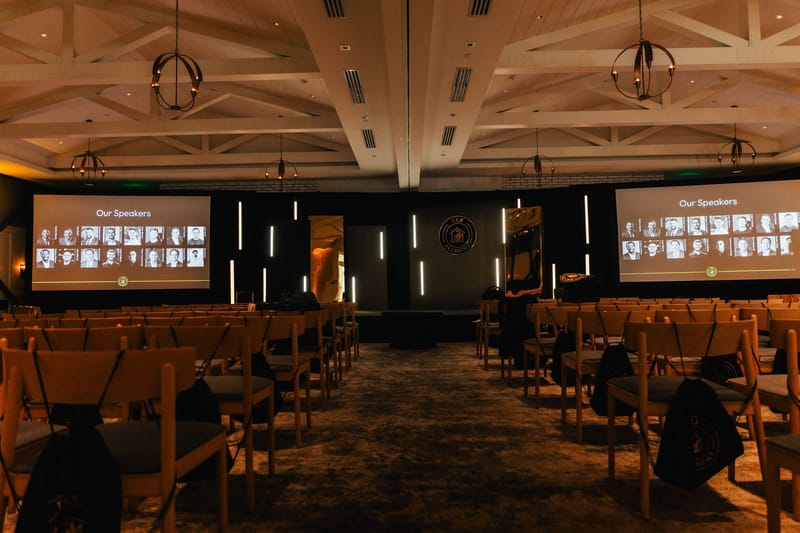 Wide view of the main room with rows of wooden chairs and screens reading Our Speakers