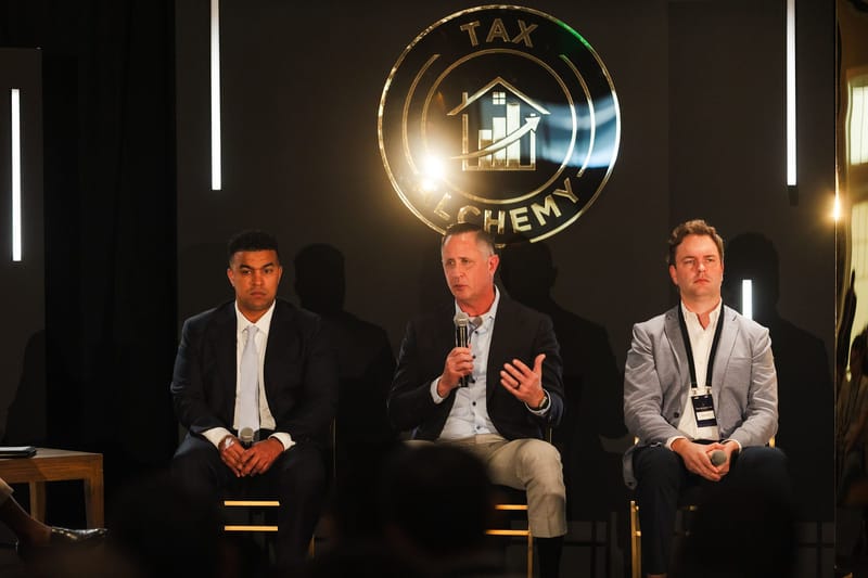 Three panelists seated on stage beneath a glowing round Tax Alchemy logo