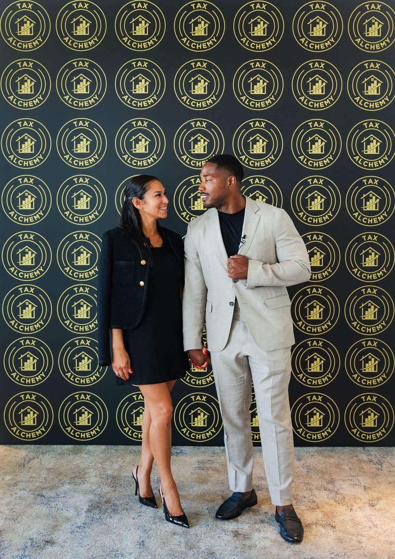 Two guests posing in front of a black Tax Alchemy logo backdrop printed with gold logos