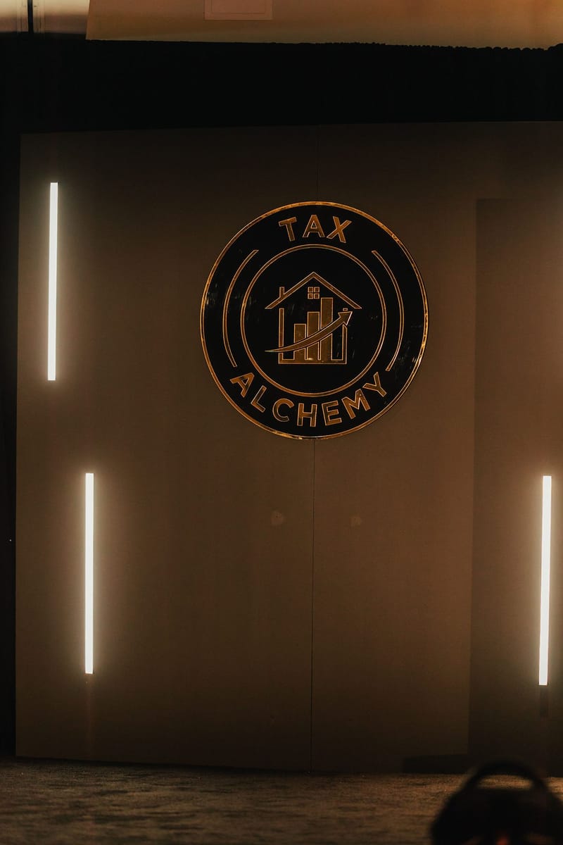 Round Tax Alchemy logo sign on a dark stage wall between vertical light bars