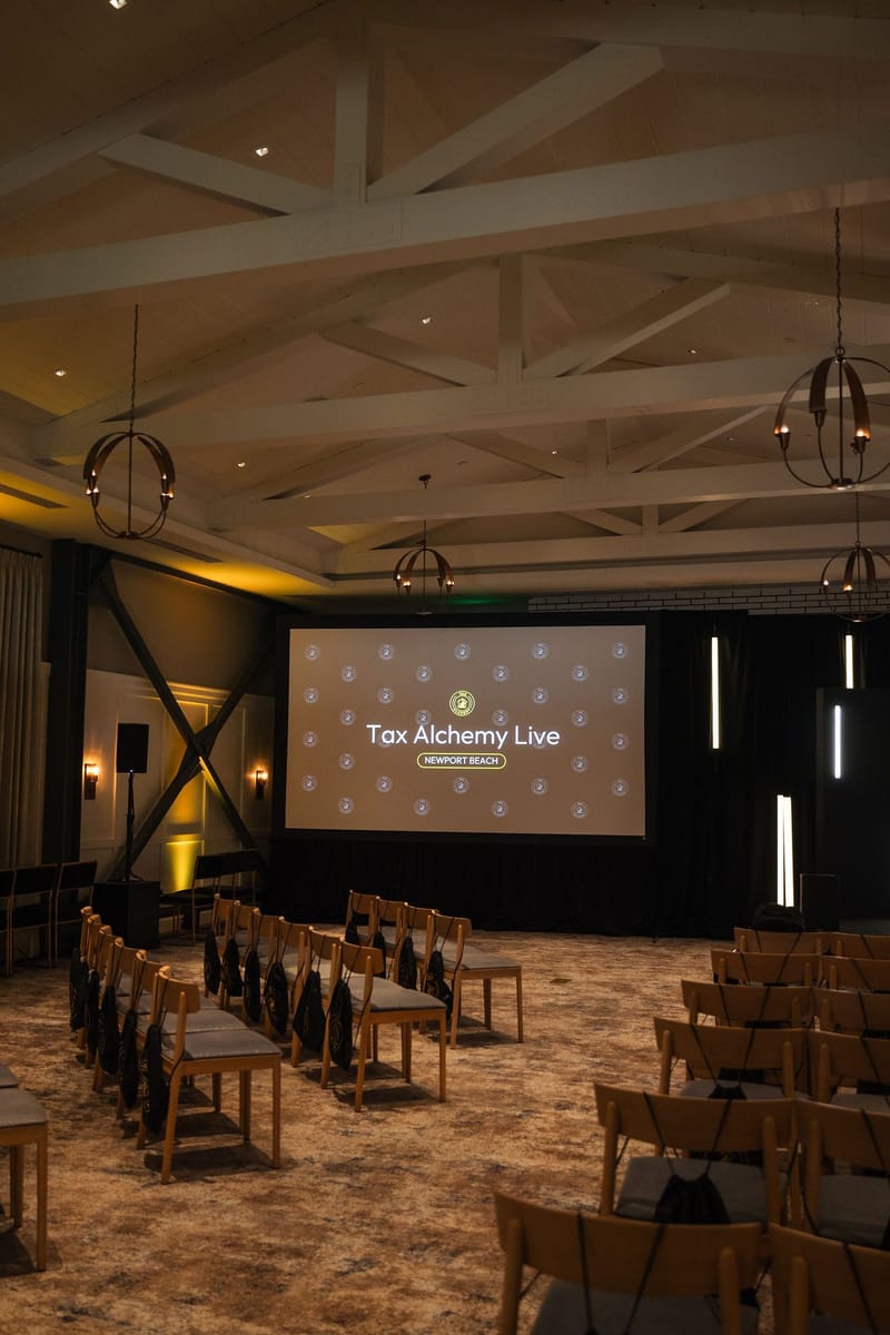 Empty room of wooden chairs under white ceiling beams, facing a Tax Alchemy Live screen