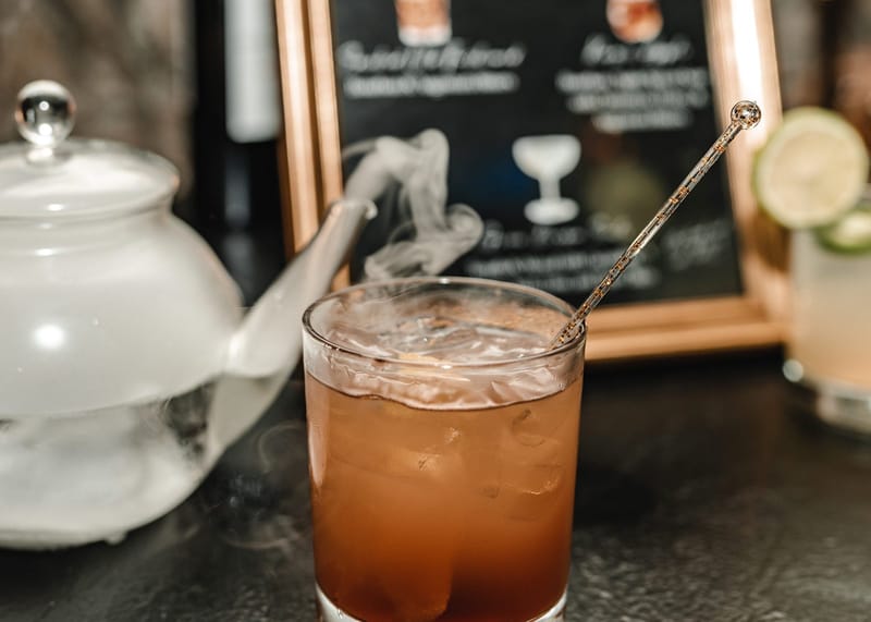 Amber cocktail with a gold stirrer in front of a fogging glass teapot and a framed drinks menu