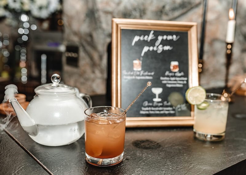 Framed pick your poison drinks menu behind a fogging teapot, an amber cocktail and a drink with a lime wheel
