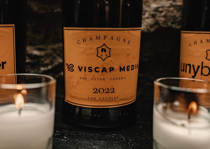 Champagne bottle with a custom Viscap Media label reading Pop. Clink. Cheers. 2022, between two candles