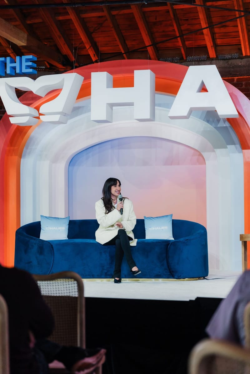 Speaker with a microphone seated on a navy velvet sofa on The Whalies stage