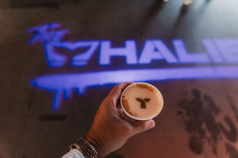 Hand holding a coffee with a whale tail design above a Whalies logo projected on the floor