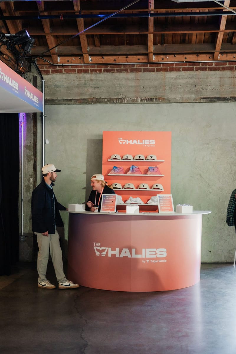 Pink Whalies merch counter with shelves of caps as two people talk