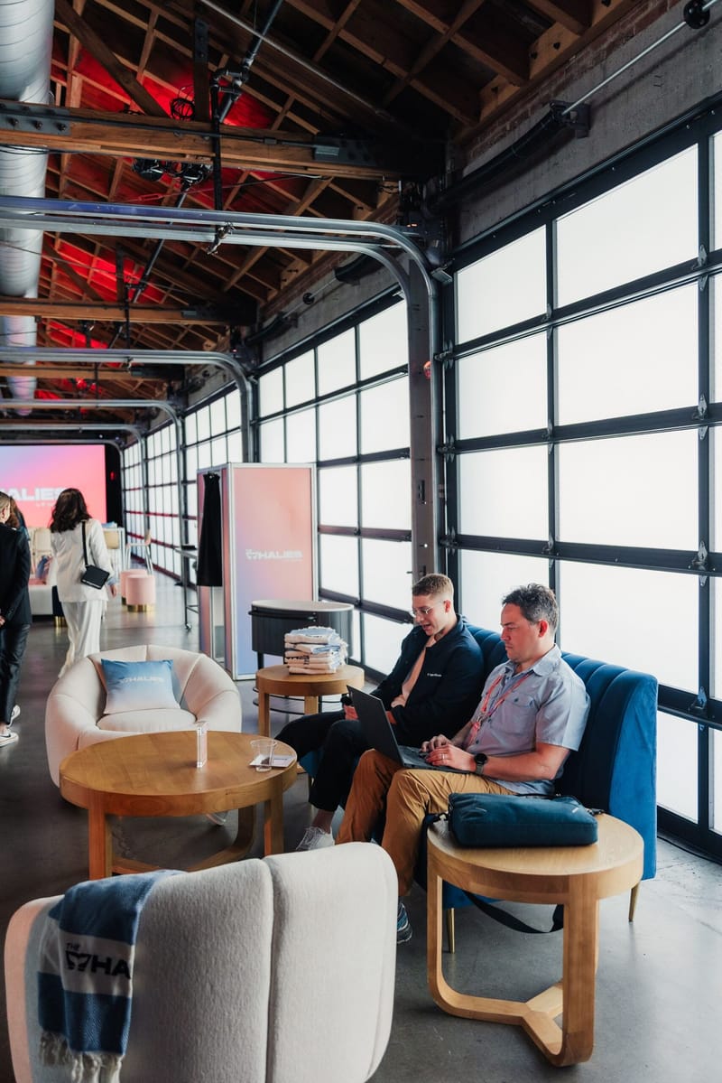 Guests on laptops in a lounge with blue armchairs and round wooden tables beside glass garage doors