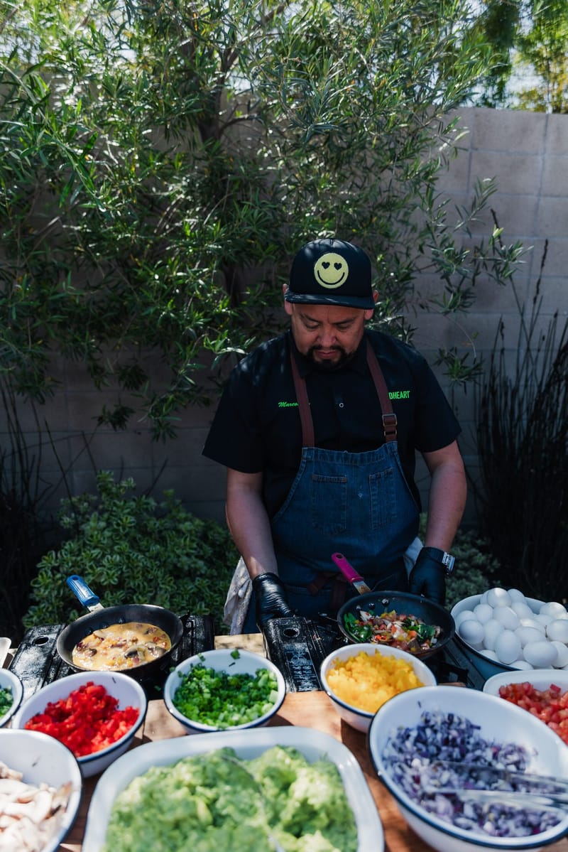 Chef in an apron at an outdoor cooking station with bowls of eggs, peppers and fresh toppings