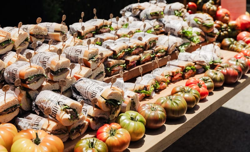 Long table of paper-wrapped sandwiches lined with heirloom tomatoes