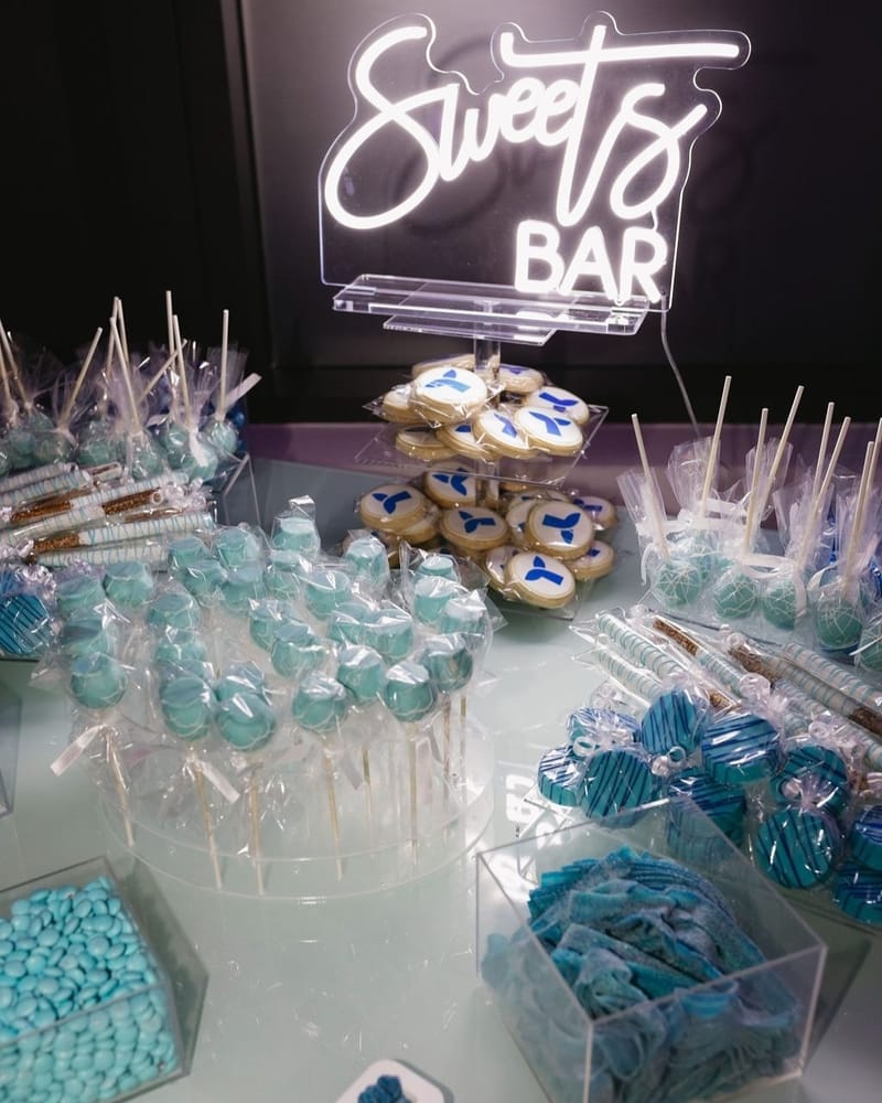 Neon Sweets Bar sign above whale tail logo cookies, blue cake pops and wrapped chocolate-dipped treats