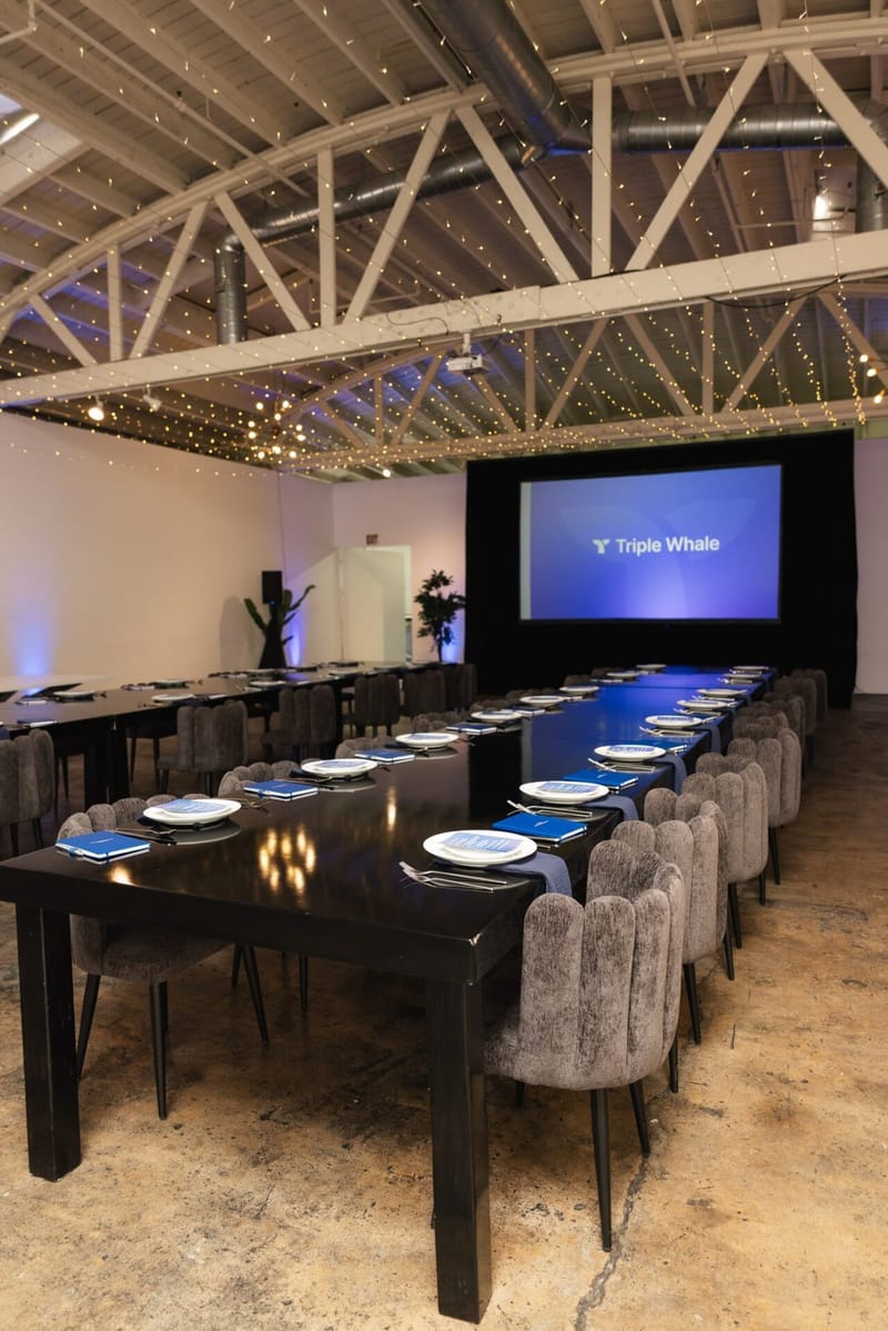 Long black table set with white plates and blue notebooks leading to a Triple Whale presentation screen