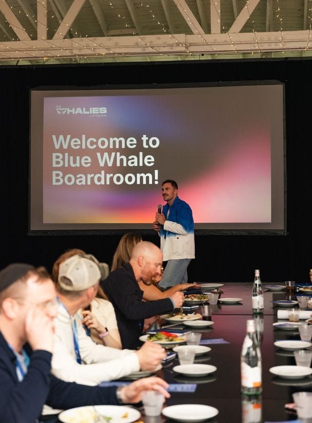 Speaker with a microphone beside a screen reading Welcome to Blue Whale Boardroom! as guests eat lunch