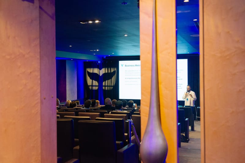 Speaker presenting to an audience in cinema seats, with a whale tail lit on the curtains, seen between wooden pillars