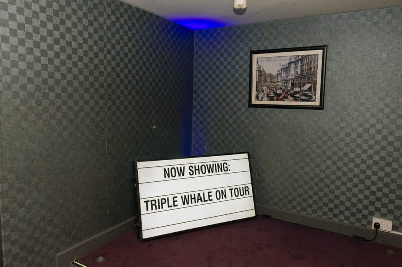 Letter board reading Now Showing: Triple Whale On Tour in the corner of a room with a framed print