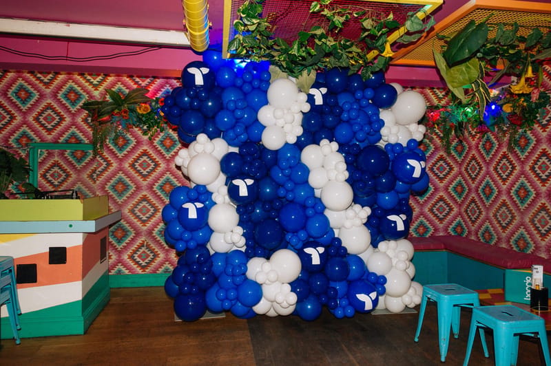 Wall of blue and white balloons with Triple Whale logos beside patterned wallpaper and teal stools