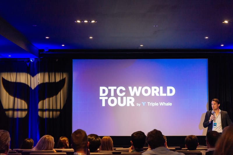 Speaker beside a cinema screen reading DTC World Tour by Triple Whale in front of a seated audience