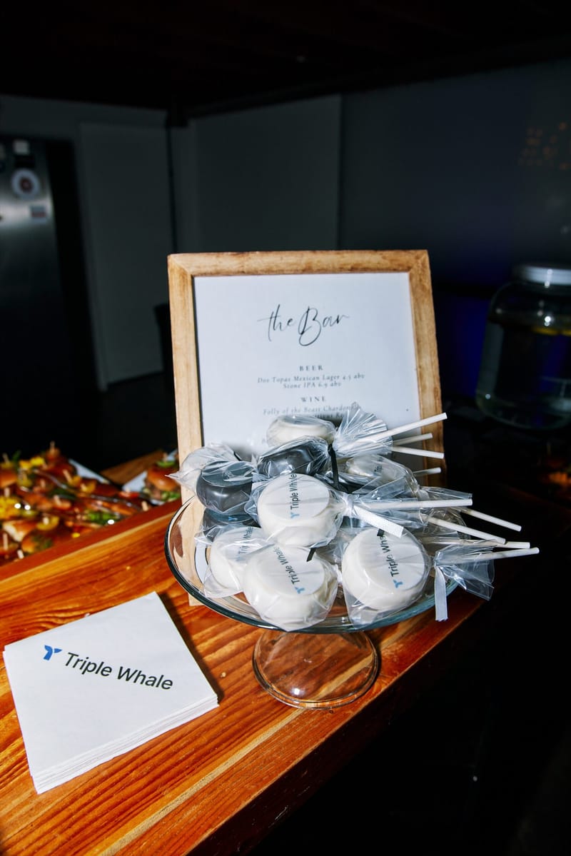 Wrapped treats on sticks printed with the Triple Whale logo in front of a framed The Bar menu