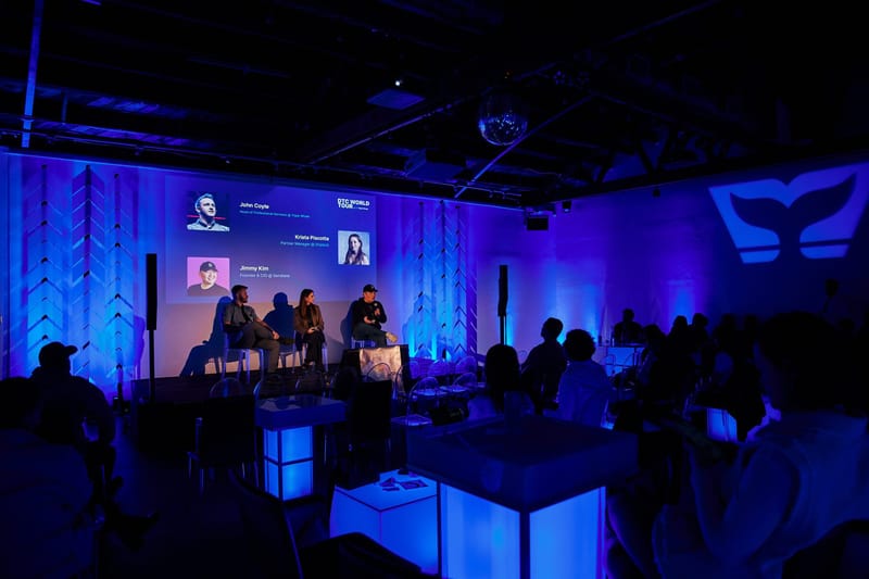 Three panelists on stage beside white chevron columns, with a whale tail logo projected on the side wall