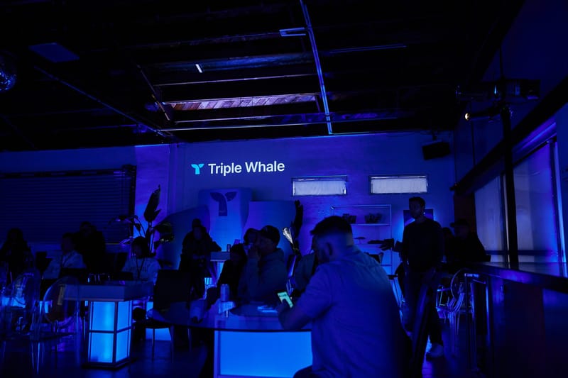Guests seated at glowing cube tables and clear chairs below Triple Whale lettering on the back wall
