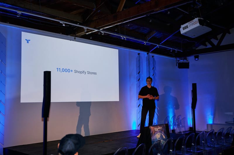 Speaker on stage beside a screen reading 11,000+ Shopify Stores, with rows of clear chairs