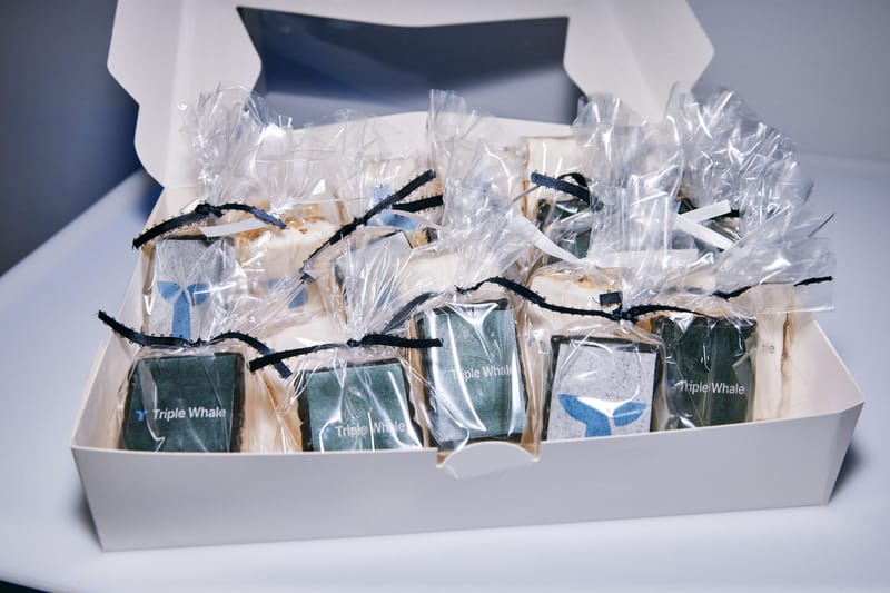 Open white box of individually wrapped treats tied with black ribbon, topped with Triple Whale logos