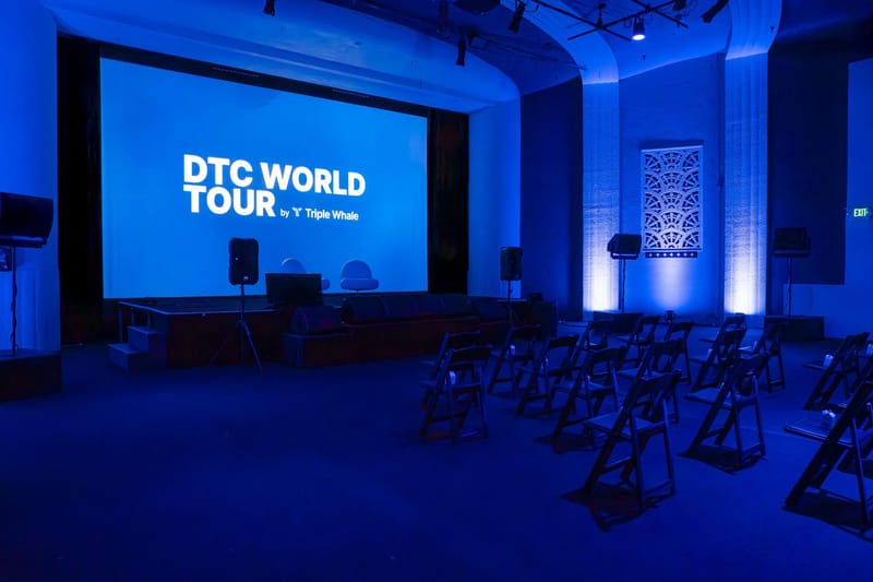 Empty blue-lit room with rows of black folding chairs facing a DTC World Tour screen