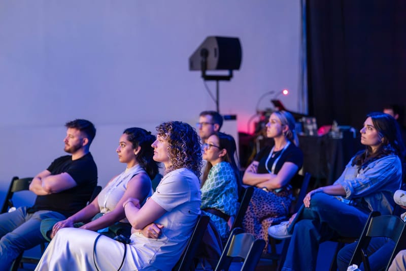 Audience seated in a row of folding chairs watching the stage under purple and blue light