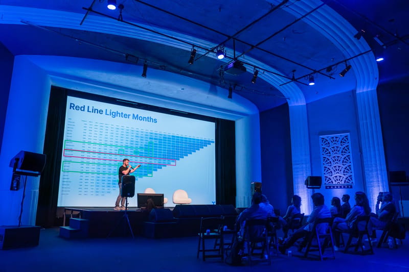 Speaker pointing at a data table slide titled Red Line Lighter Months on a large stage screen