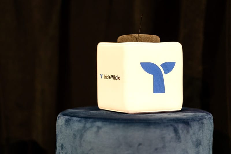 White cube microphone printed with the Triple Whale logo resting on a blue velvet pouf