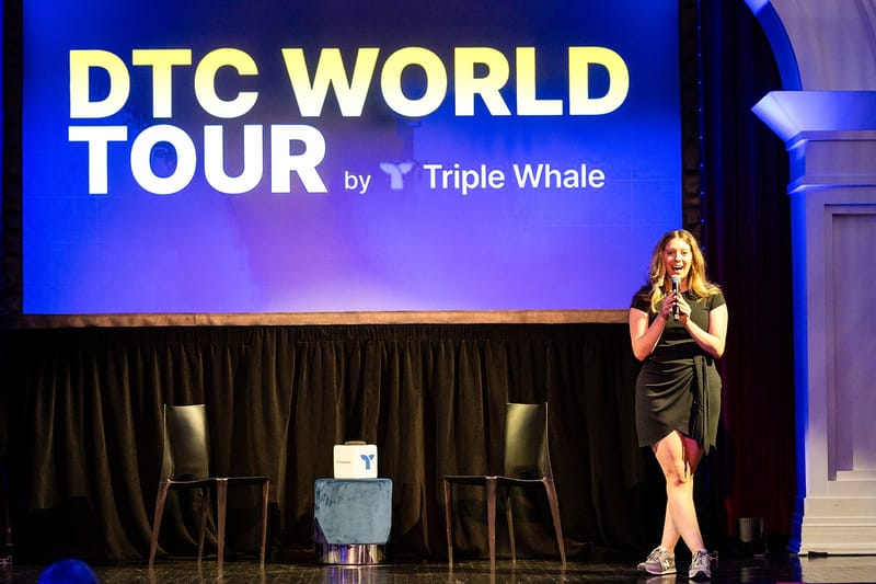 Speaker with a microphone beside a screen reading DTC World Tour by Triple Whale, with two black chairs behind