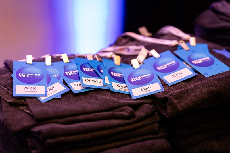 Light blue DTC World Tour ID tags printed with first names and companies, laid on stacks of dark shirts