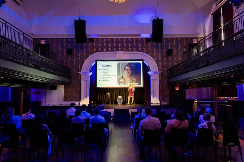 Speaker in front of an Agenda slide on stage, facing a seated audience in a hall with balconies