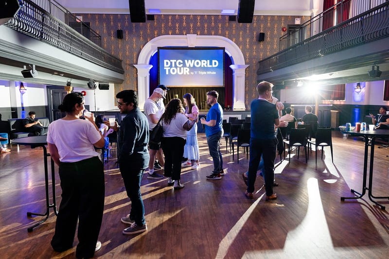 Guests talking in small groups on a wooden floor, with a DTC World Tour screen on the stage