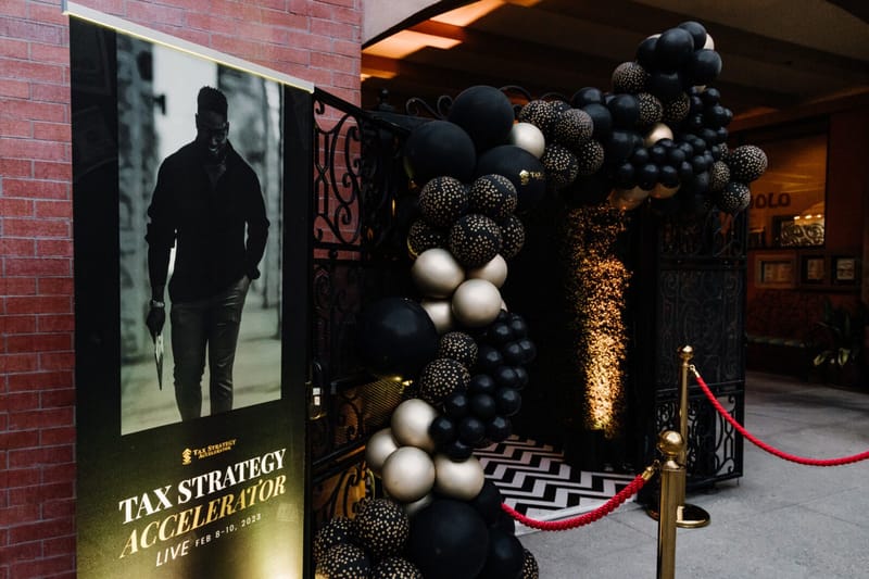 Tax Strategy Accelerator Live banner beside an entrance framed by black, gold and cream balloons and a red rope