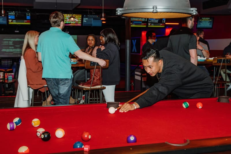 A guest lining up a shot on a red pool table while others gather at high tables