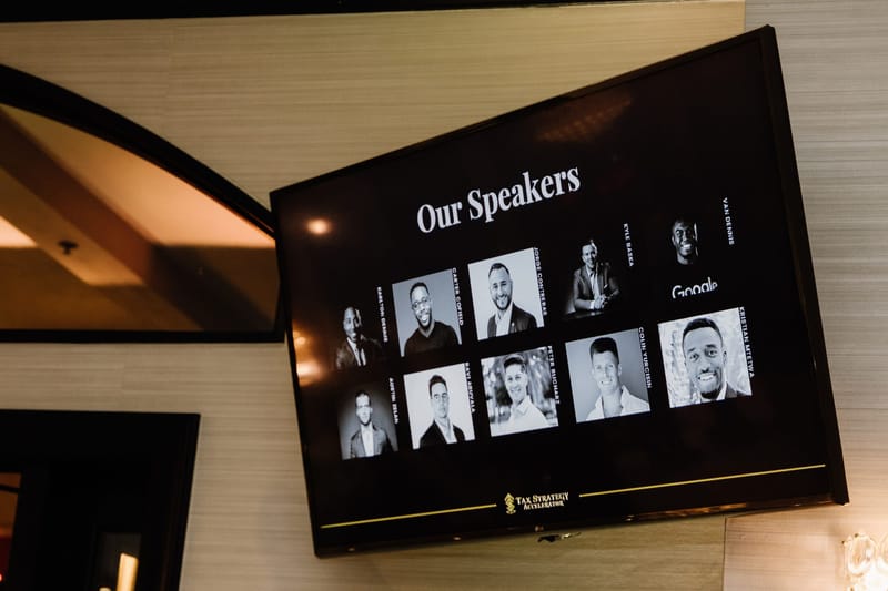 Wall-mounted screen titled Our Speakers with black and white headshots
