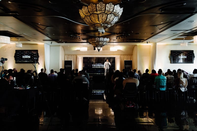 Wide view of a packed ballroom with chandeliers and a speaker on a lit stage
