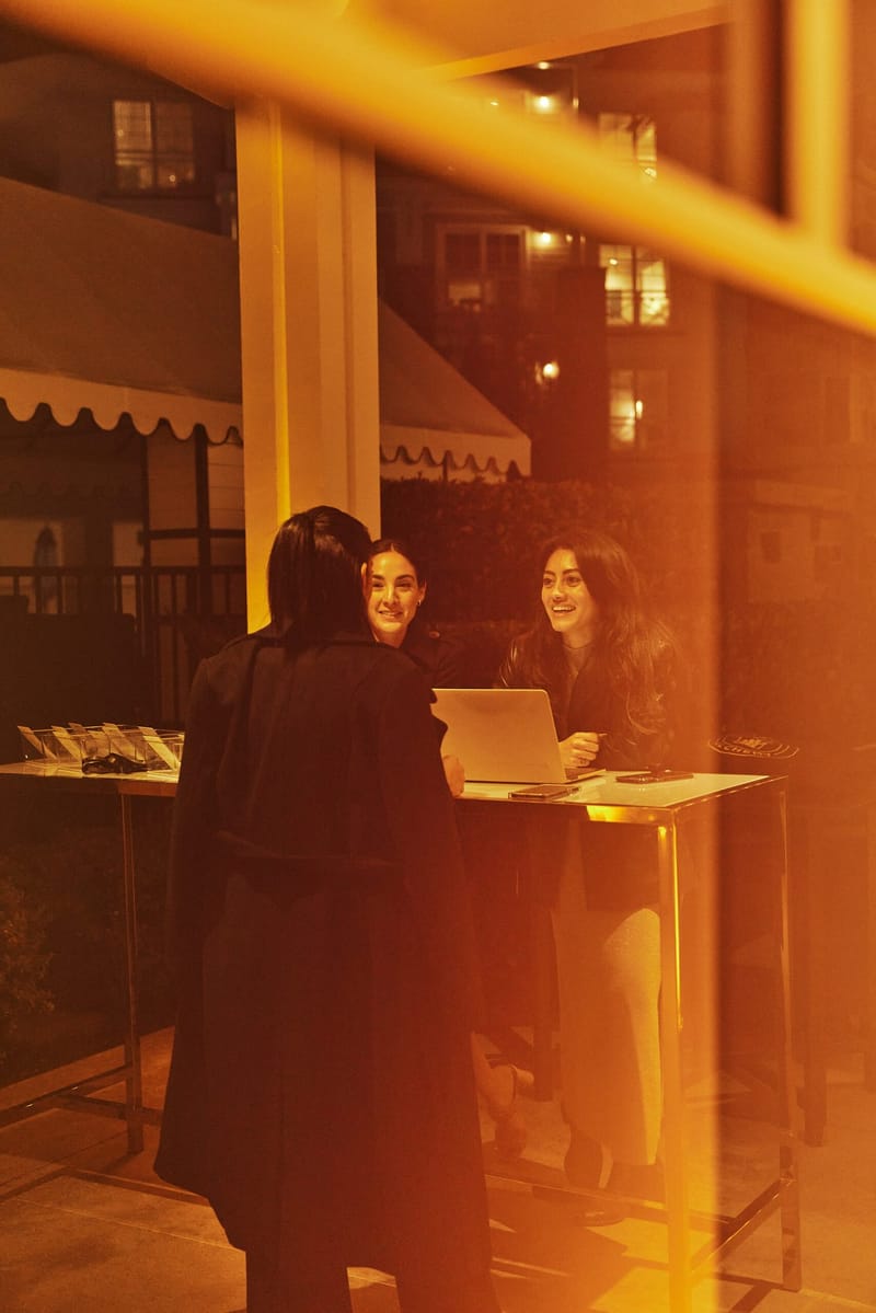 Two women at a high table with a laptop and badge holders greeting a guest, seen through glass