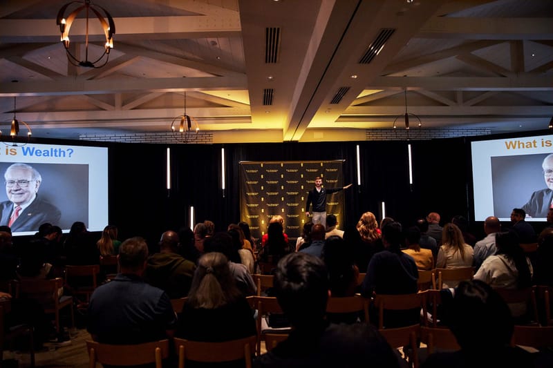 Speaker on stage before a Tax Strategy Accelerator logo backdrop, with What is Wealth? slides on two screens
