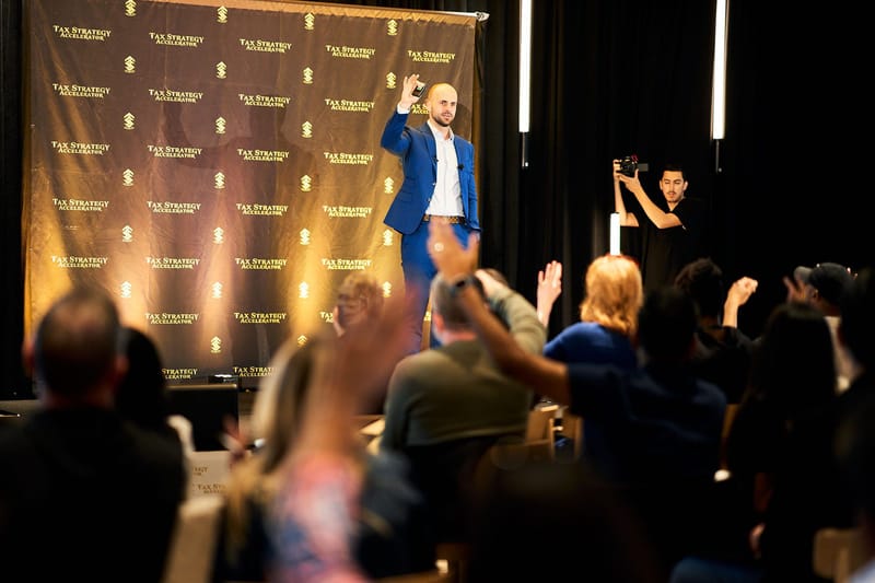 Speaker raising a fist on stage as audience members raise their hands, with a camera operator filming