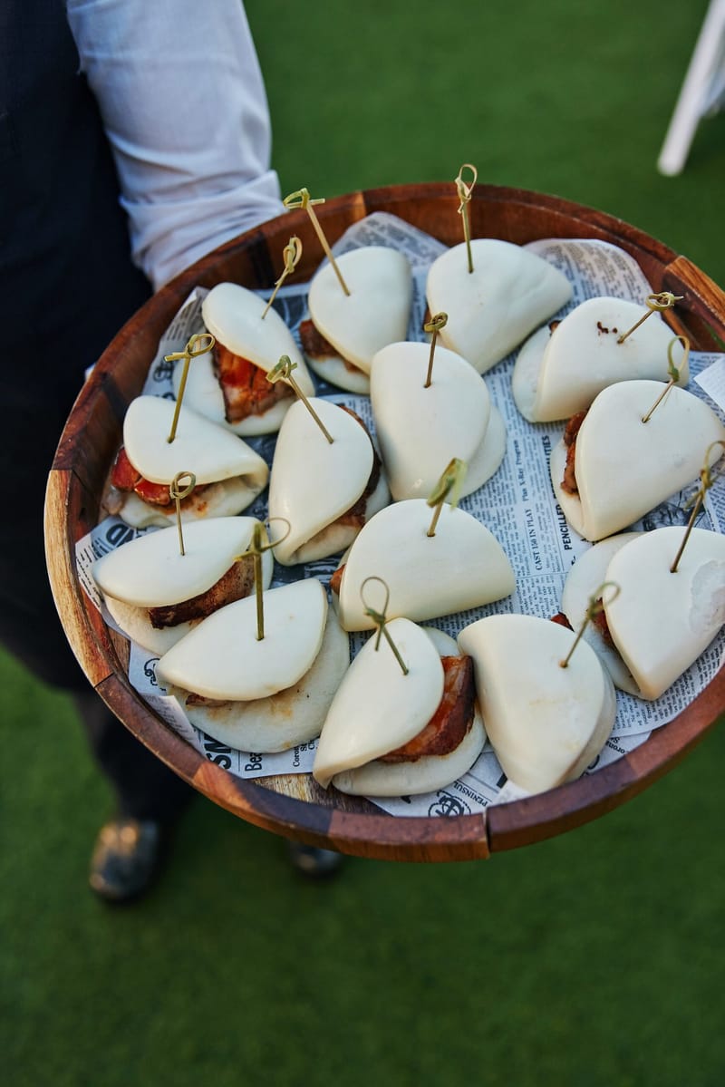Server holding a wooden bowl of bao buns with pick skewers over a lawn