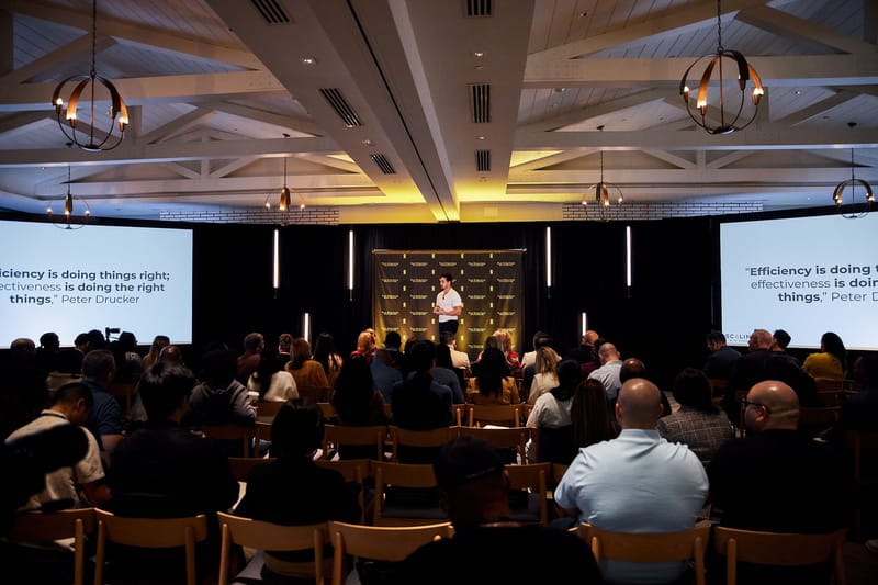 Wide view of the audience facing a speaker, with a Peter Drucker quote on both screens