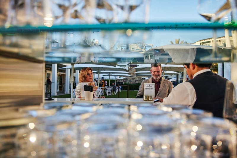 Two guests at an outdoor bar seen through rows of glasses as a bartender serves them