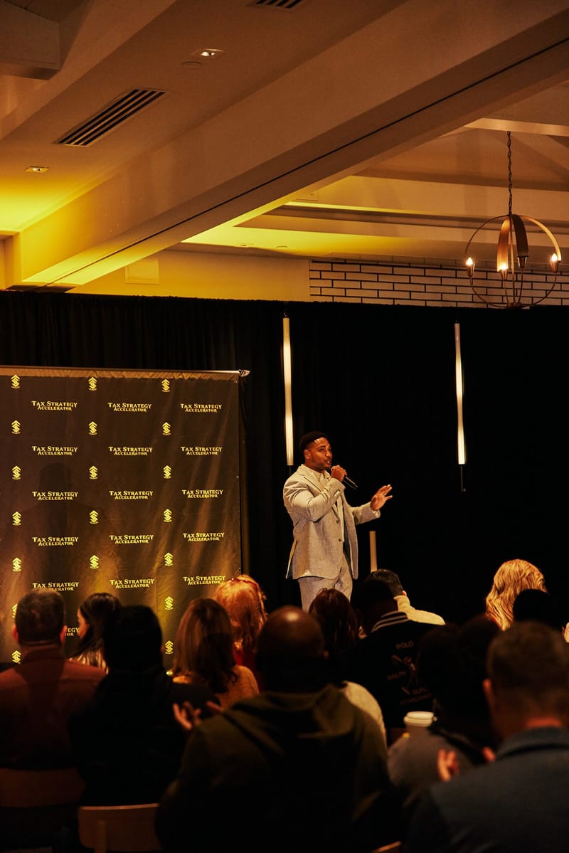 Speaker in a white suit with a microphone addressing the audience beside a logo backdrop