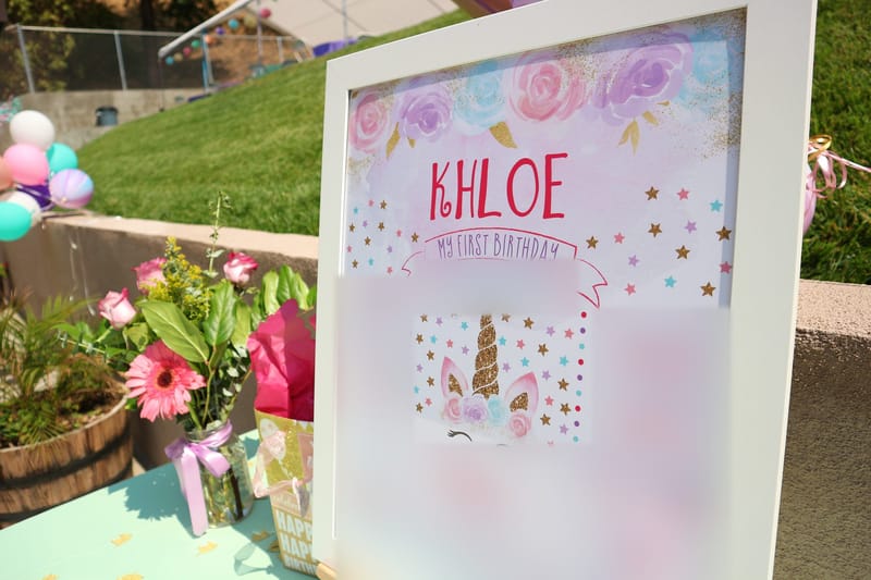 Framed unicorn first birthday board reading KHLOE on a table outdoors, with flowers and a grassy slope behind
