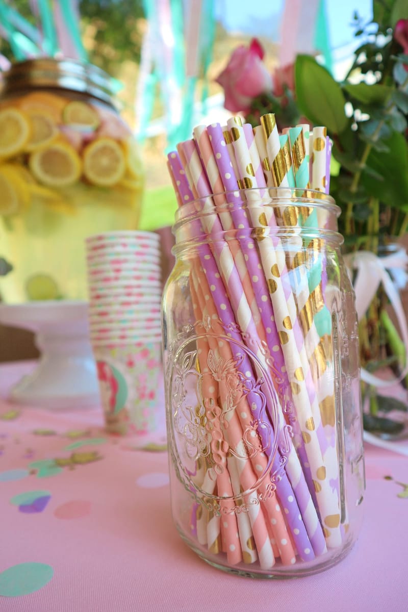 Mason jar of pastel and gold striped paper straws beside a lemon drink dispenser and paper cups